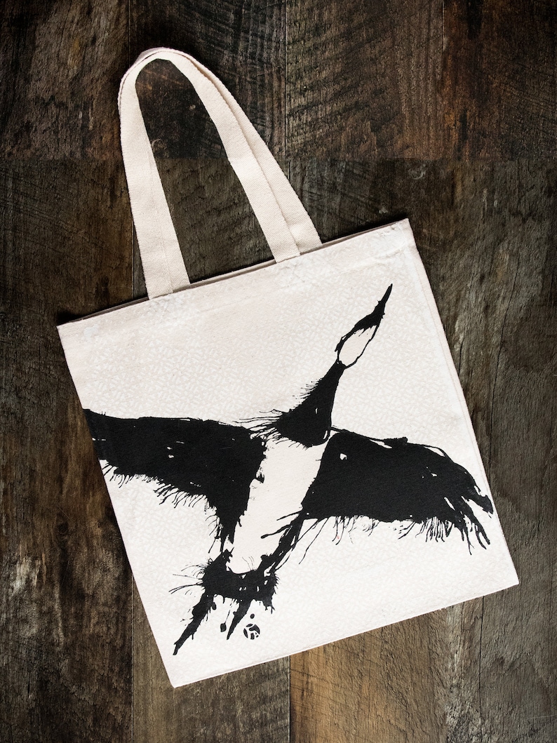 May include: A white canvas tote bag with black ink illustration of a bird in flight. The bird is stylized with a long neck and wings spread wide.