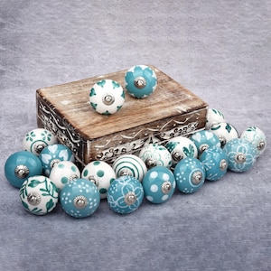 以下が含まれることがあります： 様々なデザインと色の装飾的なセラミックキャビネットノブのコレクション。ノブは、白とティール色の花柄、水玉模様、ストライプなどのパターンが特徴です。一部のノブは、彫刻が施された木製の箱の上に置かれています。