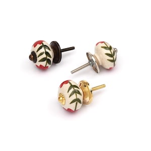 May include: Three round ceramic cabinet knobs with a cream-colored base, featuring hand-painted red floral patterns and green leaf designs. Each knob includes a metal bolt and a circular metal base plate in varying bronze and gold-toned finishes.