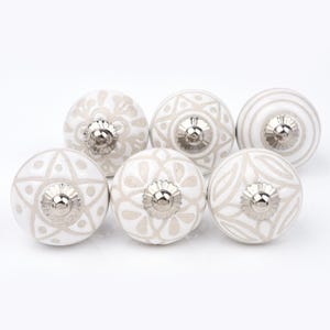 Op de afbeelding: Een set van zes ronde keramische kastknoppen met een witte basis en lichtbeige patronen. Elke knop heeft een metalen zilverkleurig middendeel met een geschulpte rand en toont diverse geometrische en bloemachtige ontwerpen.