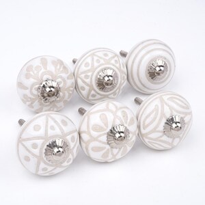 Op de afbeelding: Een set van zes ronde keramische kastknoppen met witte oppervlakken en lichtbeige patronen en ontwerpen. Elke knop heeft een centraal metalen zilverkleurig onderdeel met een geribbelde rand en een metalen schroefdraad aan de achterzijde.