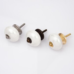 Peut inclure: Trois boutons de placard ronds en c&eacute;ramique blanche avec des bases m&eacute;talliques et des vis filet&eacute;es. Le bouton de gauche a une base argent&eacute;e, celui du centre une base bronze fonc&eacute; et celui de droite une base dor&eacute;e polie avec une vis assortie.