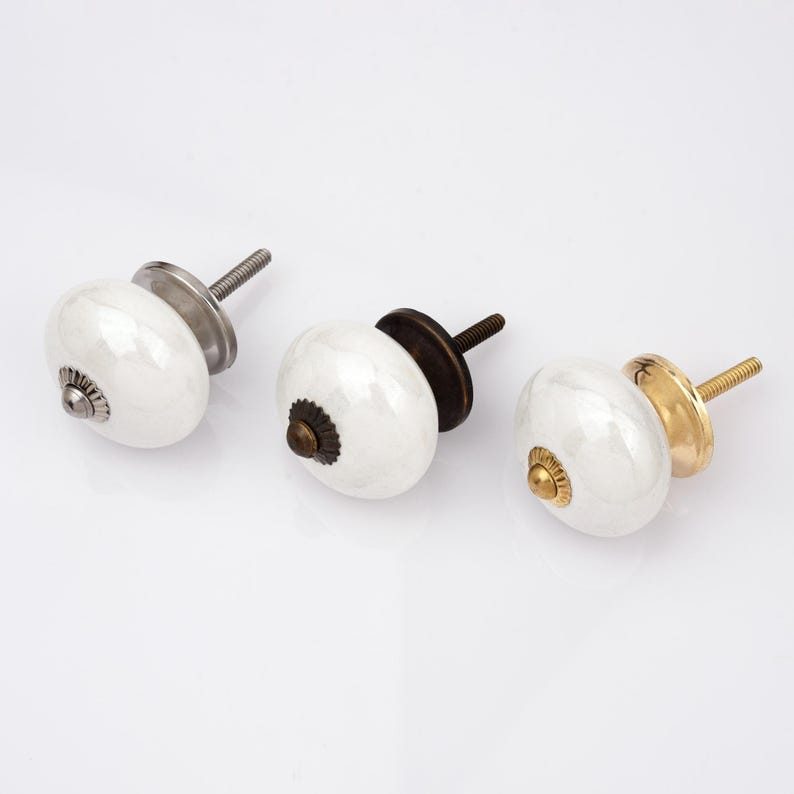 Peut inclure: Trois boutons de placard ronds en c&eacute;ramique blanche avec des bases m&eacute;talliques et des vis filet&eacute;es. Le bouton de gauche a une base argent&eacute;e, celui du centre une base bronze fonc&eacute; et celui de droite une base dor&eacute;e polie avec une vis assortie.