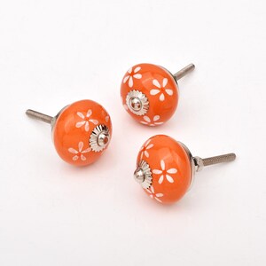 Può includere: Tre pomelli rotondi in ceramica arancione con un motivo floreale bianco. Ogni pomello presenta una base in metallo color argento e un perno filettato che sporge dal retro, progettato per il fissaggio su mobili o ferramenta.