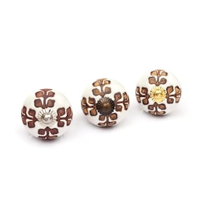 May include: Three round ceramic cabinet knobs with a white base and a repeating brown leaf-like pattern. Each knob features a central metal hardware piece, with one silver-toned, one dark bronze-toned, and one gold-toned finish on the front.