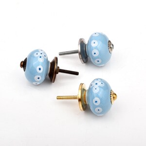 May include: Three round ceramic knobs in a light blue finish, each featuring a hand-painted white floral pattern. The knobs are equipped with metal hardware, including threaded bolts and circular base plates in bronze, silver, and gold tones.