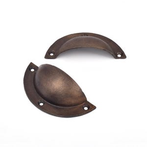 May include: Two semi-circular cup drawer pulls with a dark bronze finish. Each handle features a curved grip area and a flat mounting flange with two pre-drilled screw holes on each side, showcasing a weathered, textured metallic surface.