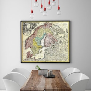 Op de afbeelding: Een ingelijste vintage kaart van Scandinavië, met gedetailleerde cartografie in verschillende kleuren. De kaart wordt weergegeven op een witte muur boven een houten eettafel met witte stoelen. Rode hanglampen hangen aan het plafond.