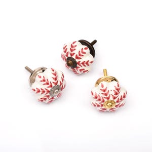 以下が含まれることがあります： スカラップエッジの白いセラミック製ノブ3個。赤い葉のような模様が描かれています。各ノブの中央には金属製のパーツがあり、背面にはネジ付きのボルトが伸びています。金具の金属仕上げはそれぞれ異なっています。