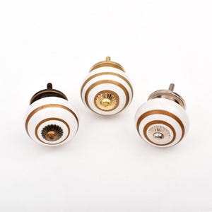 Peut inclure: Trois boutons de placard ronds en céramique avec une base blanche et des rayures concentriques dorées. Chaque bouton présente un accent métallique central et une tige filetée en métal à l'arrière pour la fixation sur des meubles.