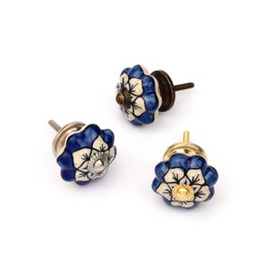 Puede incluir: Tres pomos de cerámica para cajones con forma festoneada similar a una flor y un patrón pintado en azul y blanco. Cada pomo tiene un detalle metálico central y un perno metálico roscado en la parte posterior para su instalación.
