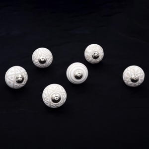 Peut inclure: Un ensemble de cinq boutons de placard ronds en céramique présentant divers motifs blancs et gris clair, notamment des designs floraux, géométriques et rayés. Chaque bouton est doté d'une petite pièce centrale en métal argenté poli.