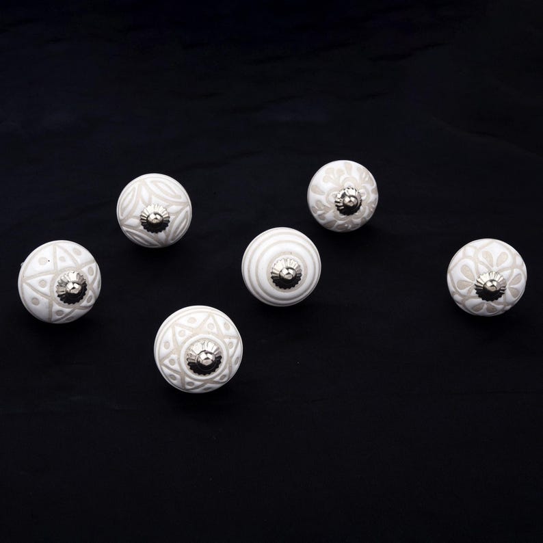Op de afbeelding: Een set van vijf ronde keramische kastknoppen met diverse witte en lichtgrijze patronen, waaronder bloemmotieven, geometrische vormen en strepen. Elke knop is afgewerkt met een klein, gepolijst zilverkleurig metalen middenstuk.
