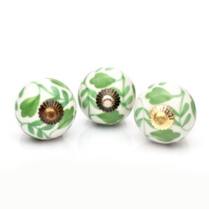 May include: Three spherical ceramic cabinet knobs featuring a white base with a hand-painted green leaf vine pattern. Each knob is topped with a metallic hardware fitting, including two with brass-toned centers and one with a gold-toned center.