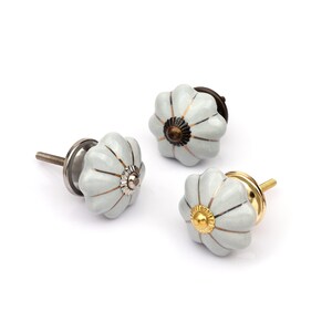May include: Three pumpkin-shaped ceramic cabinet knobs with a glossy light gray finish and vertical metallic lines. Each knob features a central decorative metal cap in different finishes, including silver, dark bronze, and polished gold tones.