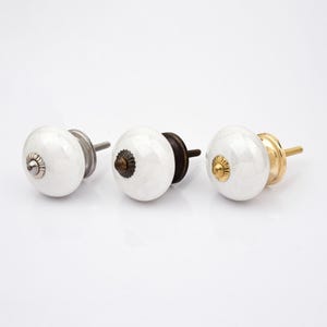 Peut inclure: Trois boutons de placard ronds en c&eacute;ramique blanche avec une finition lisse et brillante. Chaque bouton pr&eacute;sente un capuchon d&eacute;coratif central en m&eacute;tal et un boulon filet&eacute; en m&eacute;tal &agrave; l'arri&egrave;re. Les finitions m&eacute;talliques sont argent, bronze fonc&eacute; et or.