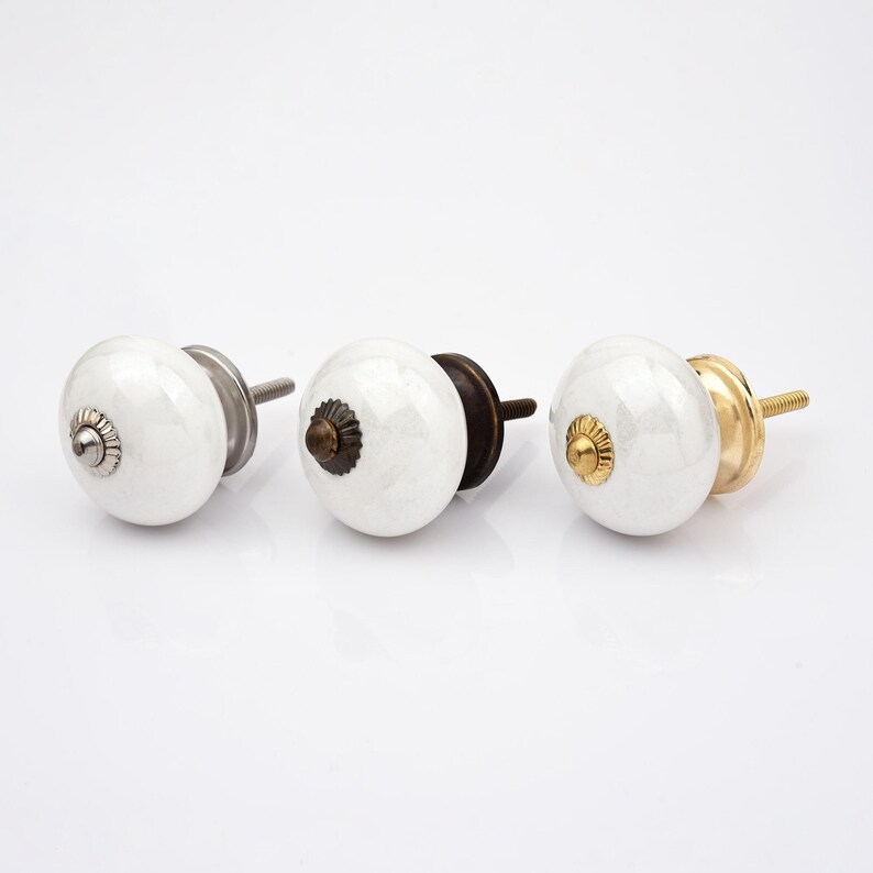 Peut inclure: Trois boutons de placard ronds en c&eacute;ramique blanche avec une finition lisse et brillante. Chaque bouton pr&eacute;sente un capuchon d&eacute;coratif central en m&eacute;tal et un boulon filet&eacute; en m&eacute;tal &agrave; l'arri&egrave;re. Les finitions m&eacute;talliques sont argent, bronze fonc&eacute; et or.