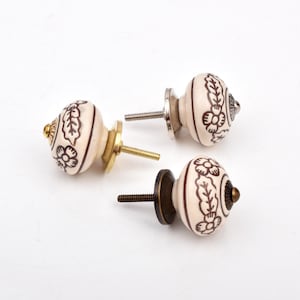 May include: Three round ceramic cabinet knobs with a cream-coloured base and brown floral patterns. Each knob features a metal mounting screw and a circular base plate in varying metallic finishes, including gold, silver, and antique bronze tones.