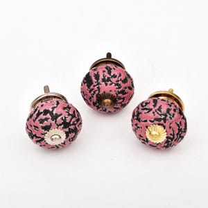 Puede incluir: Tres pomos redondos para armarios con un patrón moteado texturizado en rosa y negro. Cada pomo tiene una pieza central de metal y un tornillo roscado en la parte posterior, con acabados en tonos plata, bronce y oro.
