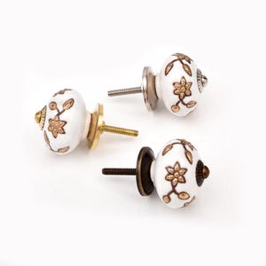 May include: Three round white ceramic cabinet knobs featuring a gold-toned floral and vine pattern. Each knob includes a threaded metal bolt and a circular base plate in varying metallic finishes, including brass, silver, and antique bronze tones.