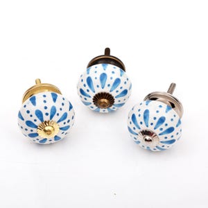 May include: Three round ceramic cabinet knobs featuring a white base with a blue painted floral pattern. Each knob has a metallic hardware base, with one gold-toned, one bronze-toned, and one silver-toned finish, each with a matching center cap.