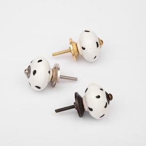 May include: Three round ceramic cabinet knobs with a white base and black spotted pattern. Each knob features a metal screw and a distinct metallic base finish, including gold, silver, and dark bronze tones, for attachment to furniture.