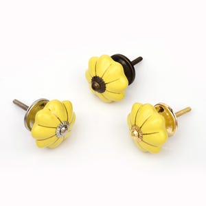May include: Three yellow ceramic drawer knobs with a pumpkin-like, fluted shape. Each knob features a central metal accent and a threaded metal bolt extending from the back. The metal hardware finishes vary between dark bronze, silver, and gold tones.