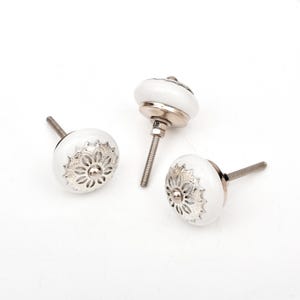 Puede incluir: Tres pomos redondos para armarios con una superficie de cerámica blanca lisa y una incrustación central de metal plateado. Cada pomo presenta un patrón decorativo de inspiración floral grabado en el metal e incluye un perno roscado.