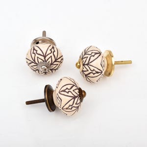May include: Three round ceramic cabinet knobs featuring a cream-colored base with a dark brown geometric floral pattern. Each knob has a metal mounting screw and a circular base plate in varying metallic finishes including silver, gold, and bronze.