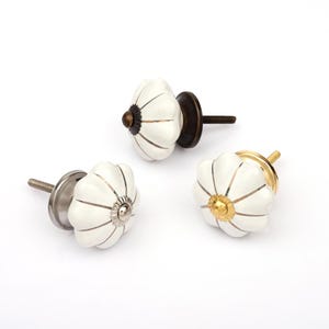 Op de afbeelding: Drie pompoenvormige kastknoppen met witte keramische oppervlakken en dunne verticale metalen lijnen. Elke knop heeft een eigen metalen voet in brons-, zilver- of goudtinten, met een schroefdraad aan de achterkant voor de installatie.