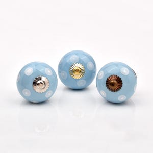 May include: Three spherical cabinet knobs in light blue with white polka dots. Each knob features a central metallic hardware attachment on the front, with varying finishes including silver, gold, and bronze tones on the circular metal plates.