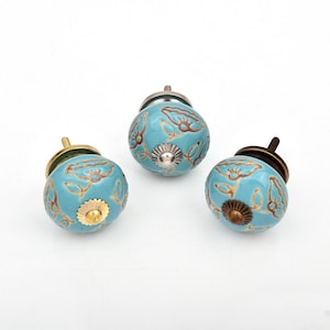 May include: Three round cabinet knobs with a light blue glazed finish and a brown floral line pattern. Each knob features a metallic center accent and a threaded metal bolt extending from the back for installation on furniture or cabinetry.