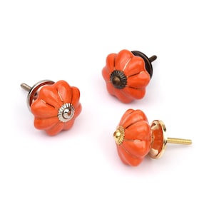 May include: Three pumpkin-shaped cabinet knobs with a glossy orange finish and fluted edges. Each knob features a central metal accent piece and a threaded metal screw post extending from the back for installation on furniture or cabinetry.