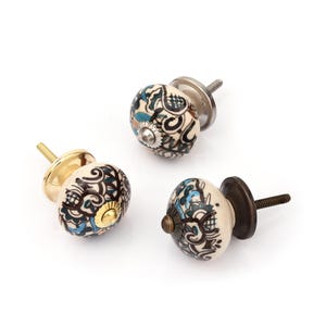 Puede incluir: Tres pomos redondos de cerámica para armarios con base color crema y patrones de remolinos en negro, azul turquesa y marrón. Cada pomo tiene una tapa central y un perno roscado de metal, con acabados en oro pulido, plata y bronce.