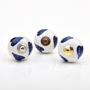 May include: Three round ceramic cabinet knobs with a white base and a blue heart pattern. Each knob features a central metallic hardware piece, with one silver-toned, one bronze-toned, and one gold-toned circular decorative cap on the front.