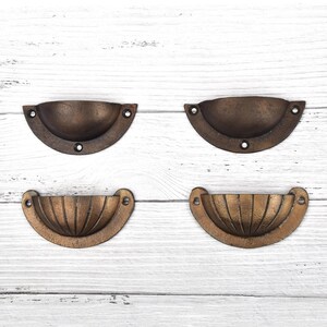 May include: Four semi-circular metal drawer pulls with a dark bronze finish. The top two pulls feature a smooth, plain surface, while the bottom two pulls have a ribbed, shell-like texture. Each pull includes three screw holes for mounting.