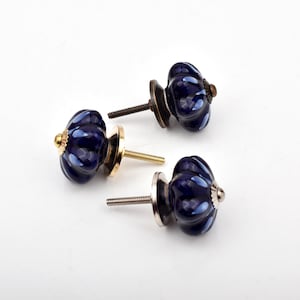 Puede incluir: Tres pomos para armarios con forma de calabaza y acabado azul oscuro brillante. Cada pomo presenta un diseño acanalado y una base metálica, con un tornillo roscado que sobresale por detrás. Los acabados varían entre latón y plateado.