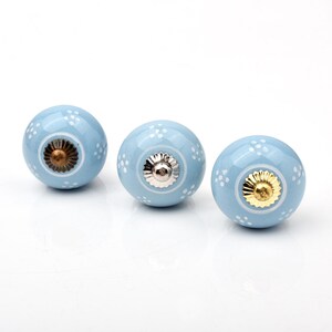 May include: Three round ceramic cabinet knobs in a light blue finish, each featuring a hand-painted white dot pattern. The center of each knob is accented with a metallic hardware piece in bronze, silver, and gold tones respectively.