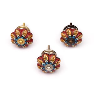 Puede incluir: Tres pomos de cerámica para cajones con un diseño en forma de flor y pétalos pintados en rojo, amarillo y azul. Cada pomo tiene un detalle metálico central y una base de tornillo roscado, con variaciones en el acabado metálico en oro y bronce.
