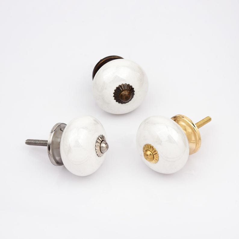Peut inclure: Trois boutons de placard ronds en c&eacute;ramique blanche avec quincaillerie m&eacute;tallique. Chaque bouton pr&eacute;sente une finition lisse et brillante avec une pi&egrave;ce centrale distincte en bronze fonc&eacute;, argent ou dor&eacute;, fix&eacute;e &agrave; une vis de montage filet&eacute;e.