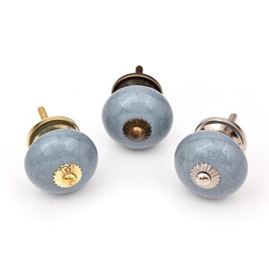 May include: Three round ceramic cabinet knobs with a light blue crackle glaze finish. Each knob features a decorative metal centre accent and a metal base. The hardware finishes include polished gold, antique bronze, and polished silver tones.