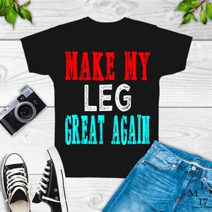 May include: Black t-shirt with the text "Make My Leg Great Again" in red, white, and blue lettering. The shirt is shown on a wooden surface with a pair of jeans and a camera.