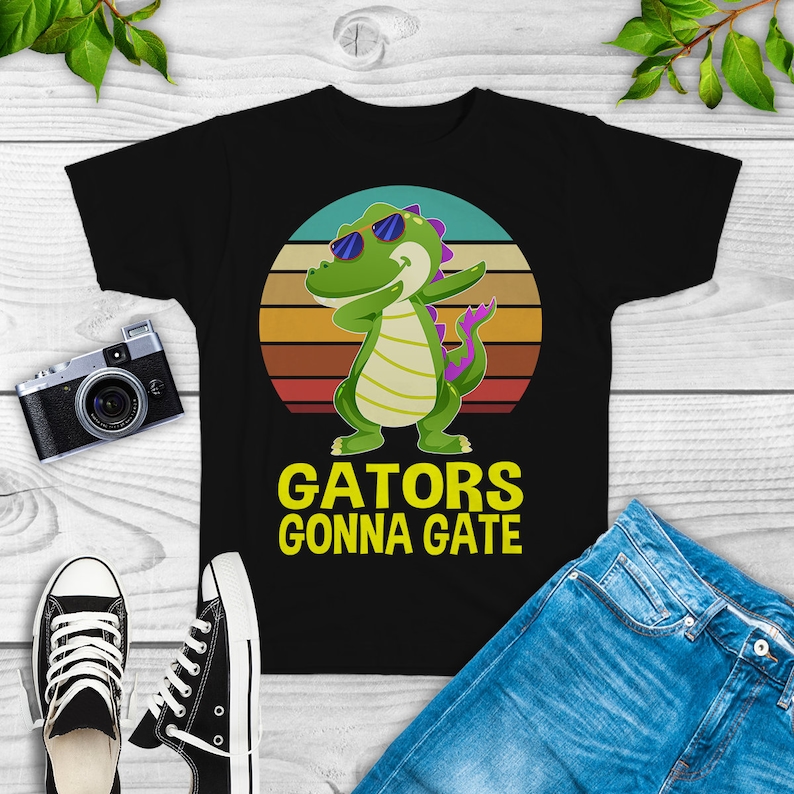 Puede incluir: Camiseta negra con un cocodrilo de dibujos animados con gafas de sol y un fondo de puesta de sol retro. El texto "GATORS GONNA GATE" est&aacute; impreso en amarillo en la camiseta.