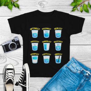 May include: Black t-shirt with a humorous graphic of glasses of water representing different perspectives. The text includes "Optimist", "Pessimist", "Realist", "Physicist", "Surrealist", "Relativist", "Utopist", "Scepticist", and "Oboist".