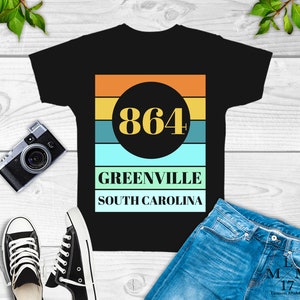 May include: Black t-shirt with a retro-style graphic design featuring a circle with the number 864 in yellow text, a multi-colored striped background, and the text "Greenville South Carolina".