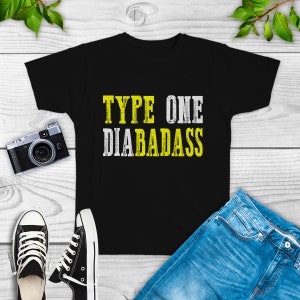 May include: Black t-shirt with the text "TYPE ONE DIABADASS" in yellow and white block letters. The shirt is displayed on a white wooden surface, alongside a pair of black and white sneakers, blue jeans, and a vintage camera.