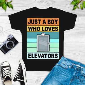 May include: Black t-shirt with a retro-style graphic that says "Just a boy who loves elevators". The graphic features a cartoon elevator with a yellow door and a blue background.