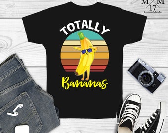 May include: Black t-shirt featuring a cartoon banana wearing sunglasses and doing a dab pose. The text "TOTALLY Bananas" is above and below the banana graphic, set against a sunset background. The shirt is displayed with jeans and sneakers.