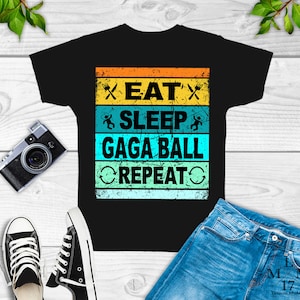May include: Black t-shirt with a vintage-style graphic that reads "Eat, Sleep, Gaga Ball, Repeat". The graphic is in a rainbow color scheme with a distressed look.