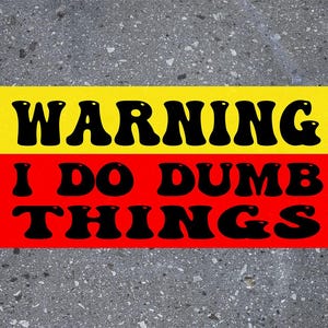 May include: A rectangular sticker with the text "WARNING I DO DUMB THINGS" in black, bold, bubble letters. The top half of the sticker is yellow, and the bottom half is red. The background is a speckled gray surface.